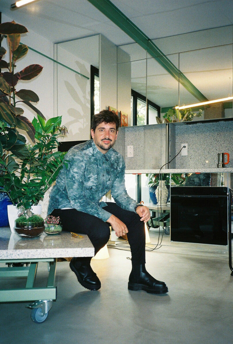 A person sits on a table in a modern room with plants, mirrors, and large windows. They wear a patterned shirt, black pants, and boots, and hold a mug.