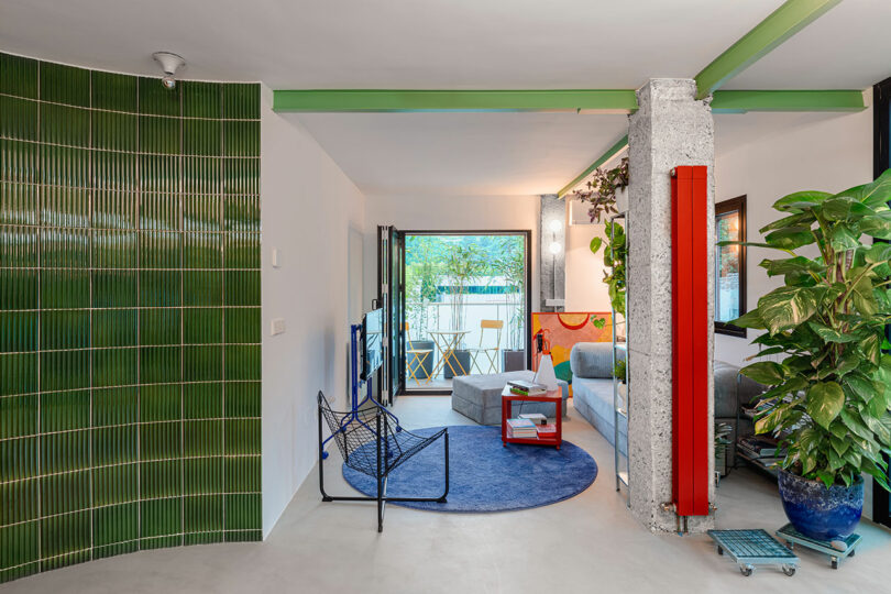 Modern living room with green-tiled accent wall, blue circular rug, and a mix of contemporary furniture. Large potted plants and a small balcony are visible in the background.