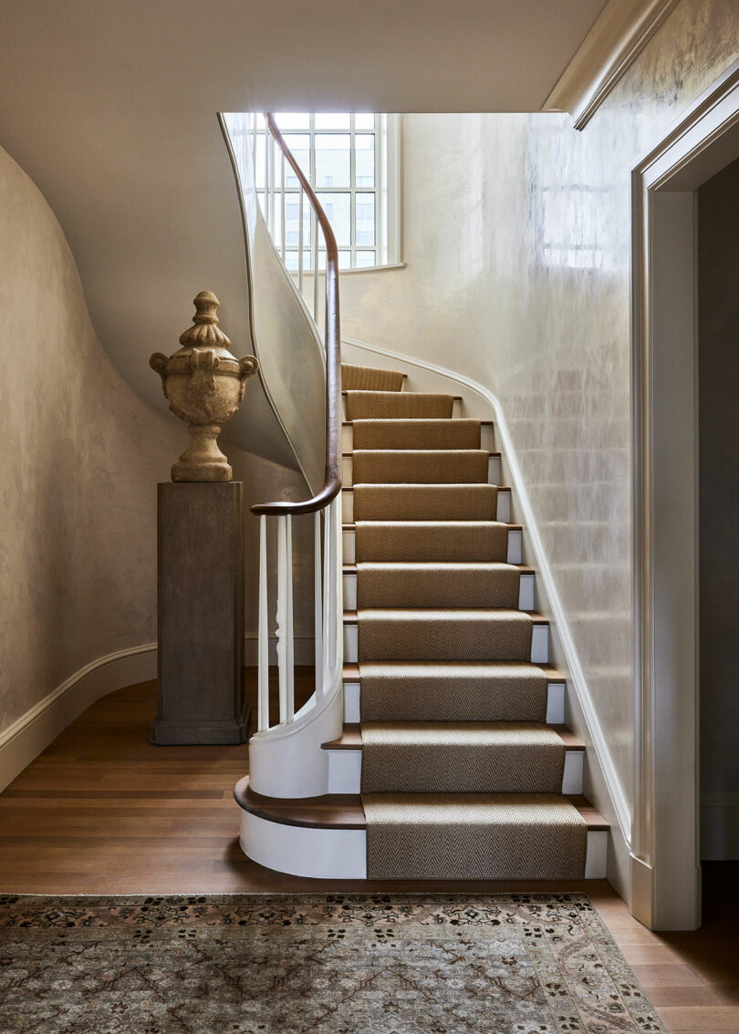 A narrow, carpeted staircase curves upwards, flanked by a white wall. A decorative urn sits on a pedestal near the base. Light filters through a window above, casting shadows reminiscent of Elizabeth Graziolo's elegant designs. A door frame is visible on the right.