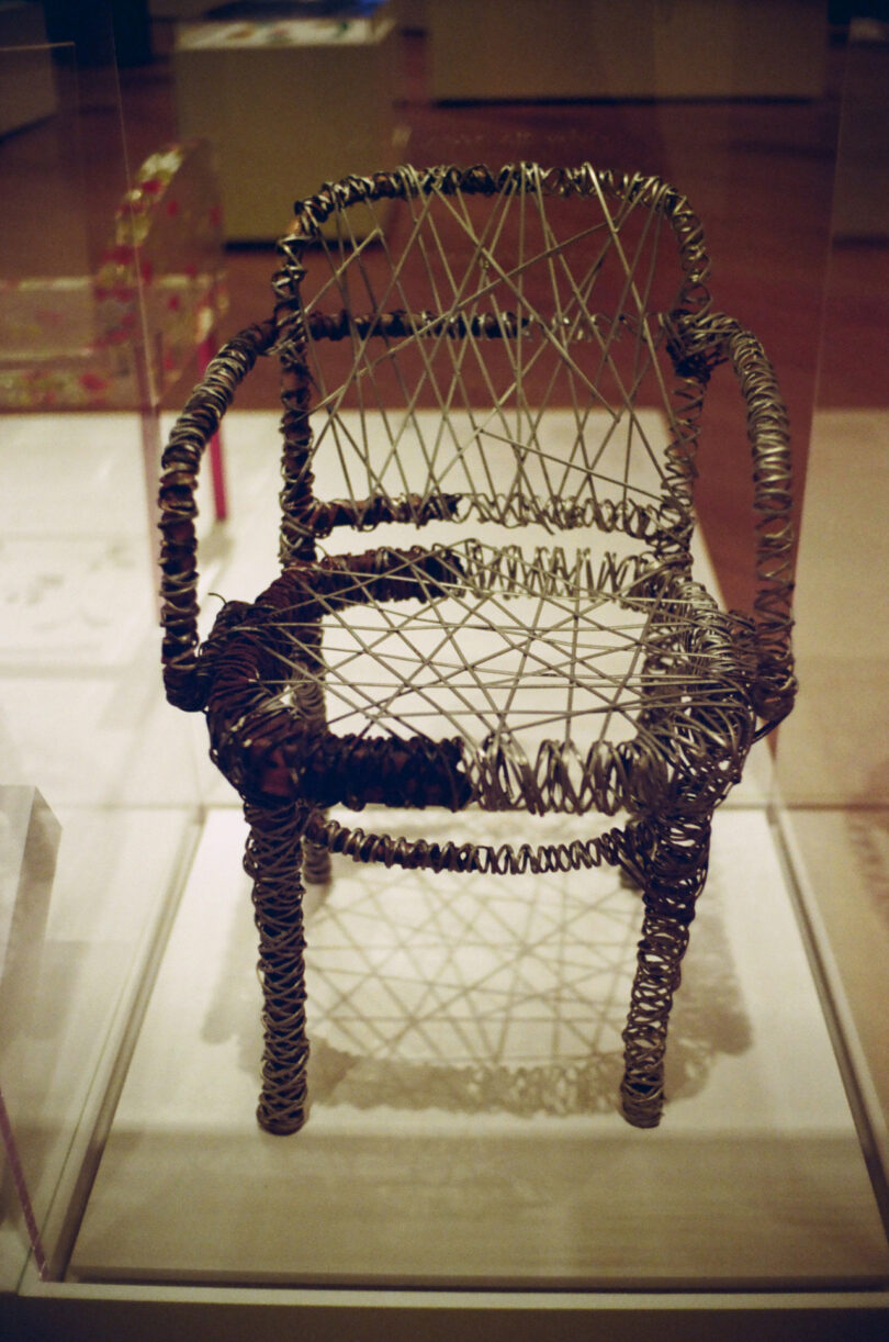 A wire chair sculpture with a complex pattern of intersecting metal strands on display in a glass case.