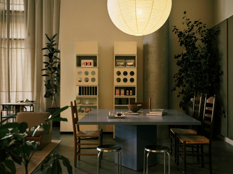 A modern dining area with a large paper lantern overhead, a blue table surrounded by wooden chairs, shelving units, and various plants.